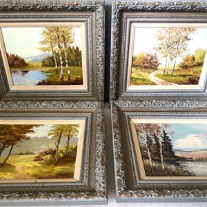 May include: Four landscape oil paintings in ornate, distressed silver-grey frames. Each piece depicts a natural scene with trees, water, or paths in muted earth tones. The frames feature intricate, raised scrollwork patterns along the edges.
