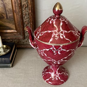 May include: A deep red lidded vessel with a pedestal base and two handles, featuring a white painted ribbon and bow pattern across the surface. The lid is topped with a gold-colored finial, and the rim and handles are accented with gold trim.