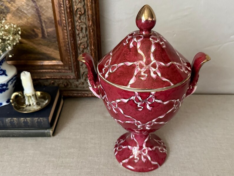 May include: A deep red lidded vessel with a pedestal base and two handles, featuring a white painted ribbon and bow pattern across the surface. The lid is topped with a gold-colored finial, and the rim and handles are accented with gold trim.