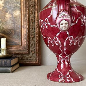 May include: A deep red ceramic urn featuring a raised white ribbon pattern and a sculpted human face relief on the upper section. The vessel has a pedestal base, a flared rim, and gold-toned trim accents along the edges and the sculpted face.