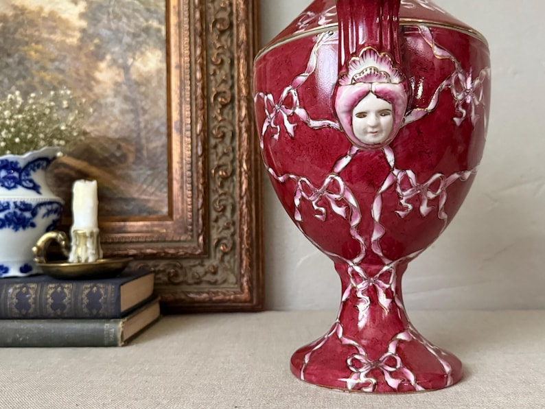 May include: A deep red ceramic urn featuring a raised white ribbon pattern and a sculpted human face relief on the upper section. The vessel has a pedestal base, a flared rim, and gold-toned trim accents along the edges and the sculpted face.
