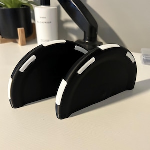 Può includere: Due supporti semicircolari in plastica nera con accenti bianchi lungo i bordi curvi. Ogni supporto presenta una base piatta e una finitura opaca liscia, progettata per mantenere gli oggetti in posizione verticale e stabile.
