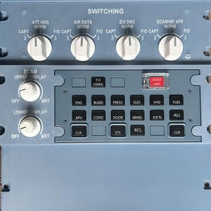 May include: A blue aircraft cockpit control panel featuring a switching section with four white rotary knobs, two display brightness knobs, and a grid of black push buttons labeled with functions like ENG, BLEED, PRESS, ELEC, HYD, FUEL, APU, COND, DOOR, WHEEL, F/CTL, ALL, CLR, STS, and RCL, plus a red EMER CANC button.