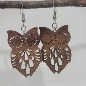 May include: A pair of brown wooden owl earrings with intricate cutouts. The earrings are suspended from silver hooks.