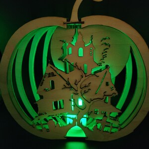 May include: A wooden pumpkin-shaped Halloween decoration with a light-up haunted house scene inside. The house is cut out and the light shines through the windows, creating a spooky glow.