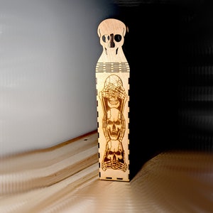 Op de afbeelding: Een houten doos met een schedelontwerp. De doos heeft drie schedels erop, elk met een ander handgebaar. De bovenste schedel bedekt zijn ogen, de middelste schedel bedekt zijn oren en de onderste schedel bedekt zijn mond.