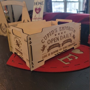 以下が含まれることがあります： 「Cupid's Emporium Open Daily」と「Love is always the secret ingredient」という文字が刻まれた木製の箱。箱にはハートとキューピッドのデザインが施されています。