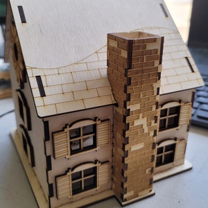 May include: A wooden model of a house with a brick chimney, two windows with shutters, and a decorative roof. The house is made of light brown wood and has a white painted roof.