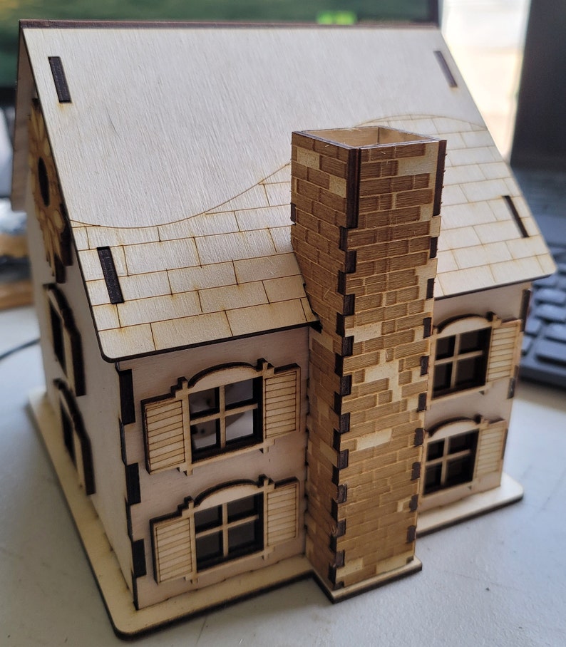 May include: A wooden model of a house with a brick chimney, two windows with shutters, and a decorative roof. The house is made of light brown wood and has a white painted roof.