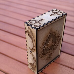 May include: A wooden box with a black border and intricate laser-cut designs. The box features a stylised ace of spades with a bird perched on top.