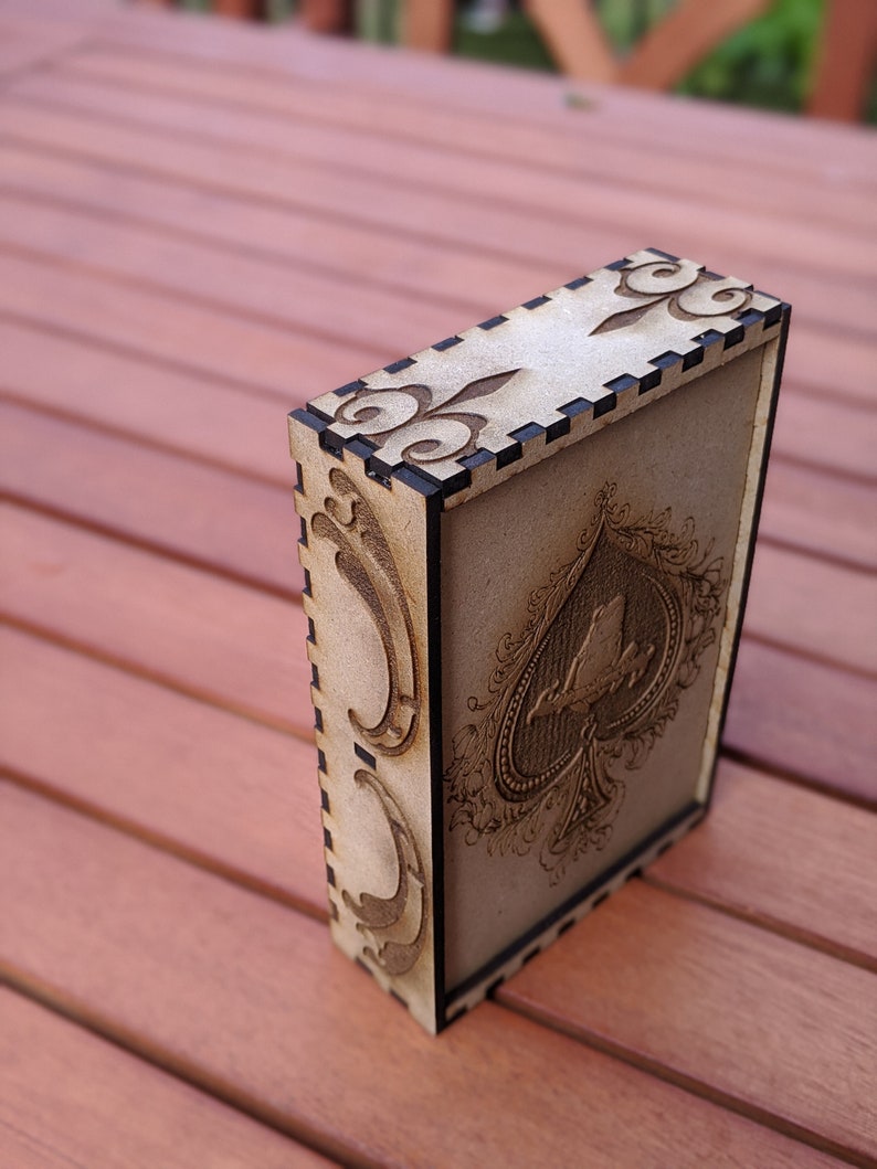 May include: A wooden box with a black border and intricate laser-cut designs. The box features a stylised ace of spades with a bird perched on top.