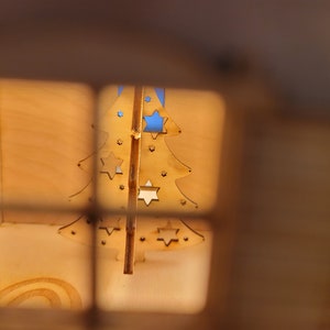 May include: A wooden Christmas tree ornament with cut-out stars, viewed through a window in a wooden house.
