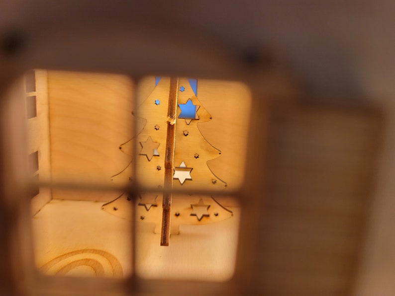 May include: A wooden Christmas tree ornament with cut-out stars, viewed through a window in a wooden house.