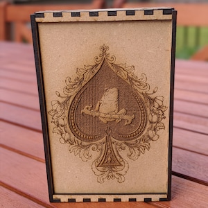 May include: A wooden box featuring a decorative design of a spade playing card with a frog sitting on it. The box is brown with black trim.