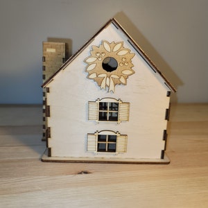 May include: A wooden Christmas house with a wreath on the front, two windows with shutters, and a chimney. The house is painted white and has a natural wood finish.