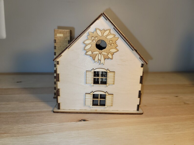 May include: A wooden Christmas house with a wreath on the front, two windows with shutters, and a chimney. The house is painted white and has a natural wood finish.