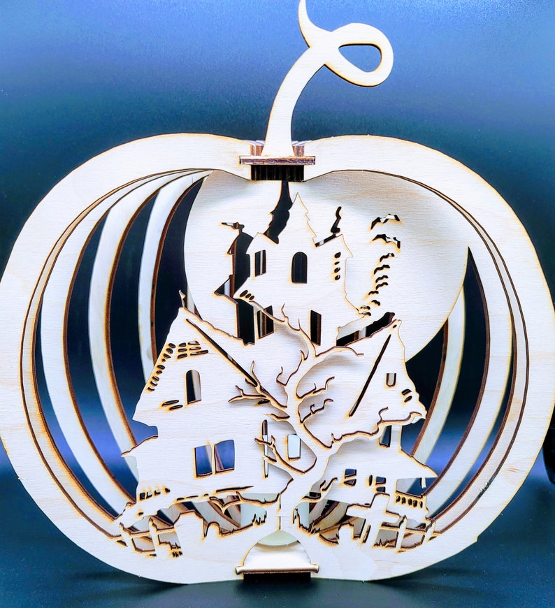 May include: A wooden pumpkin shaped ornament with a laser cut design of a spooky house and a tree. The pumpkin is made of multiple layers of wood and has a hollow center.