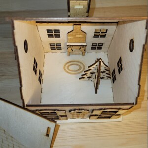 May include: A wooden Christmas house with a removable roof, revealing a fireplace, a Christmas tree, and a circular rug inside. The house has windows and a chimney.