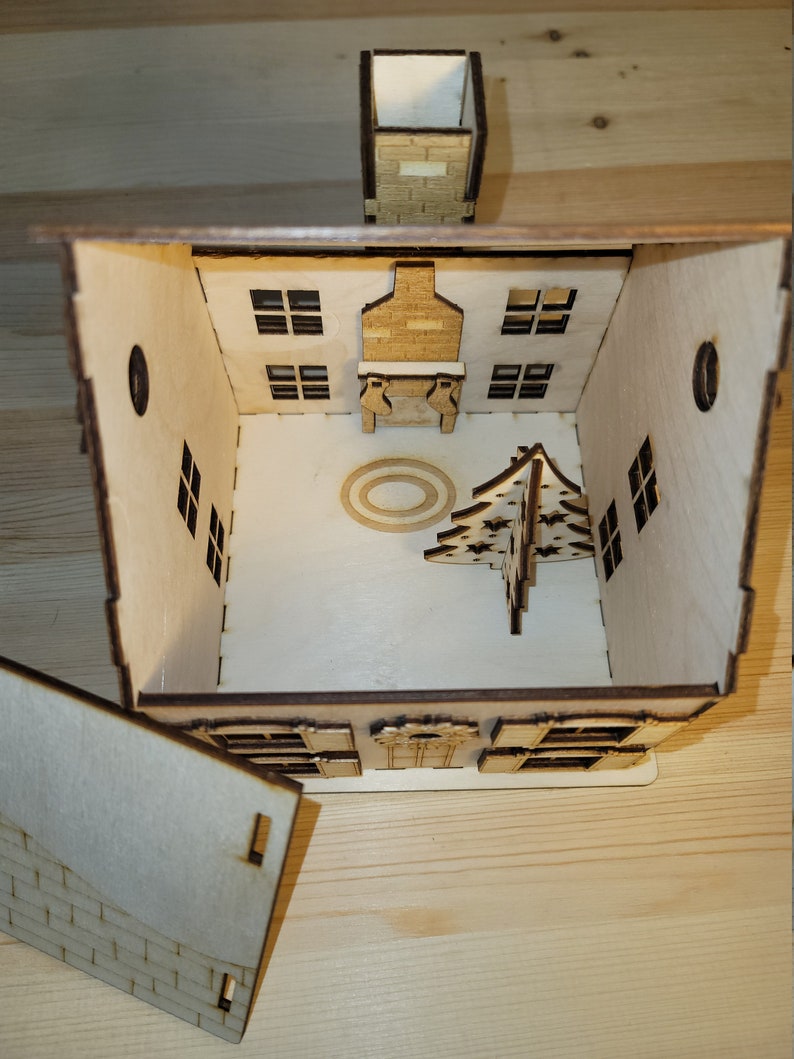 May include: A wooden Christmas house with a removable roof, revealing a fireplace, a Christmas tree, and a circular rug inside. The house has windows and a chimney.