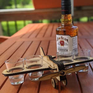 May include: Wooden aeroplane shot glass holder with four shot glasses and a bottle of Jim Beam Kentucky Straight Bourbon whiskey on a wooden table.