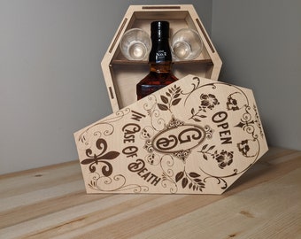 Peut inclure: Une boîte en bois en forme de cercueil avec un couvercle à charnières. Le couvercle est ouvert et présente un motif de crâne et d'os croisés avec les mots "Ouvrir en cas de décès". La boîte contient une bouteille de whisky et deux verres à shot.