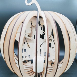 May include: A wooden pumpkin shaped lantern with a carved design of a haunted house. The lantern has a slot for a candle in the center.
