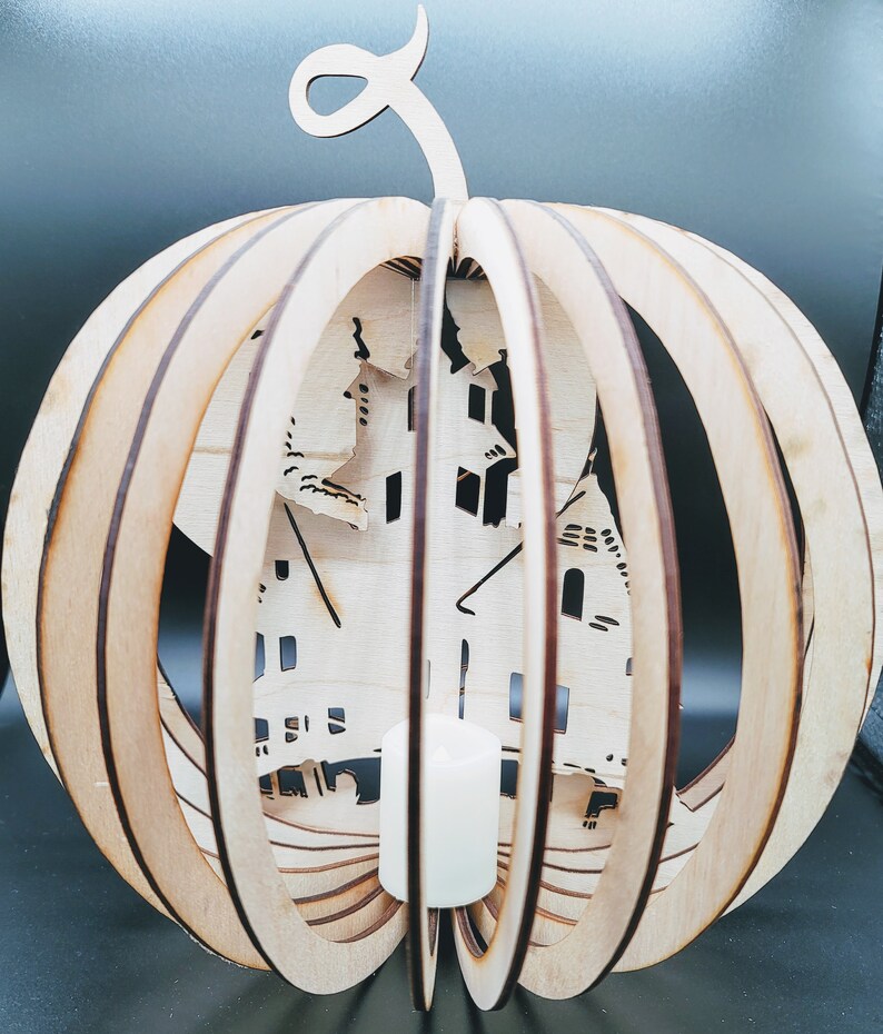 May include: A wooden pumpkin shaped lantern with a carved design of a haunted house. The lantern has a slot for a candle in the center.