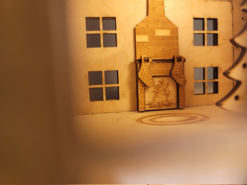May include: A wooden model of a house with a fireplace and stockings hanging from the mantle. The house has windows and a roof. The fireplace has a fire burning inside.