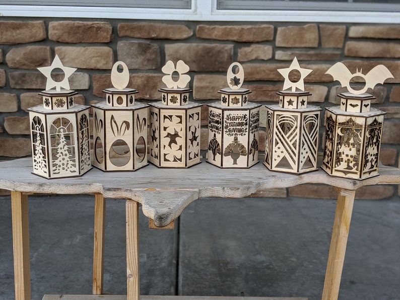 Peut inclure: Six lanternes &agrave; bougies en bois avec des motifs diff&eacute;rents. Les lanternes sont en bois et pr&eacute;sentent une vari&eacute;t&eacute; de motifs d&eacute;coup&eacute;s, notamment des &eacute;toiles, des fleurs, des lapins et une dinde. Les lanternes sont toutes de la m&ecirc;me taille et de la m&ecirc;me forme et sont dispos&eacute;es en rang&eacute;e sur une table en bois.