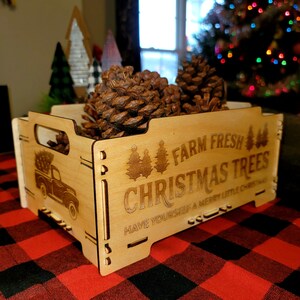 May include: A wooden box with the text "Farm Fresh Christmas Trees" and "Have Yourself a Merry Little Christmas" on the front. The box is filled with pine cones and a small truck with a Christmas tree on the back is visible on the side of the box.