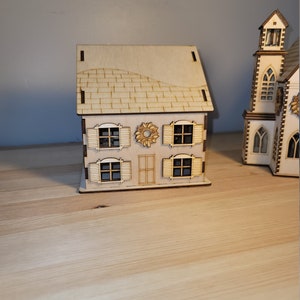 May include: A wooden model of a house with a brown roof, white walls, and windows with shutters. The house has a wreath on the door and is sitting on a wooden surface.