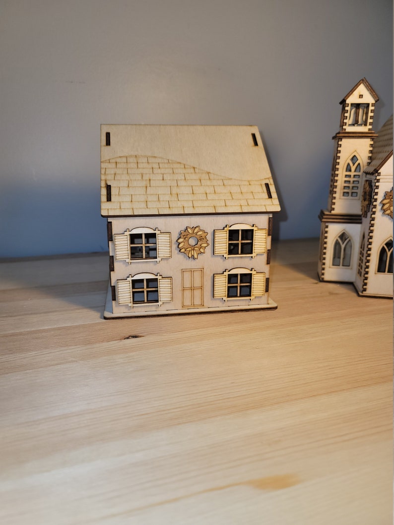 May include: A wooden model of a house with a brown roof, white walls, and windows with shutters. The house has a wreath on the door and is sitting on a wooden surface.