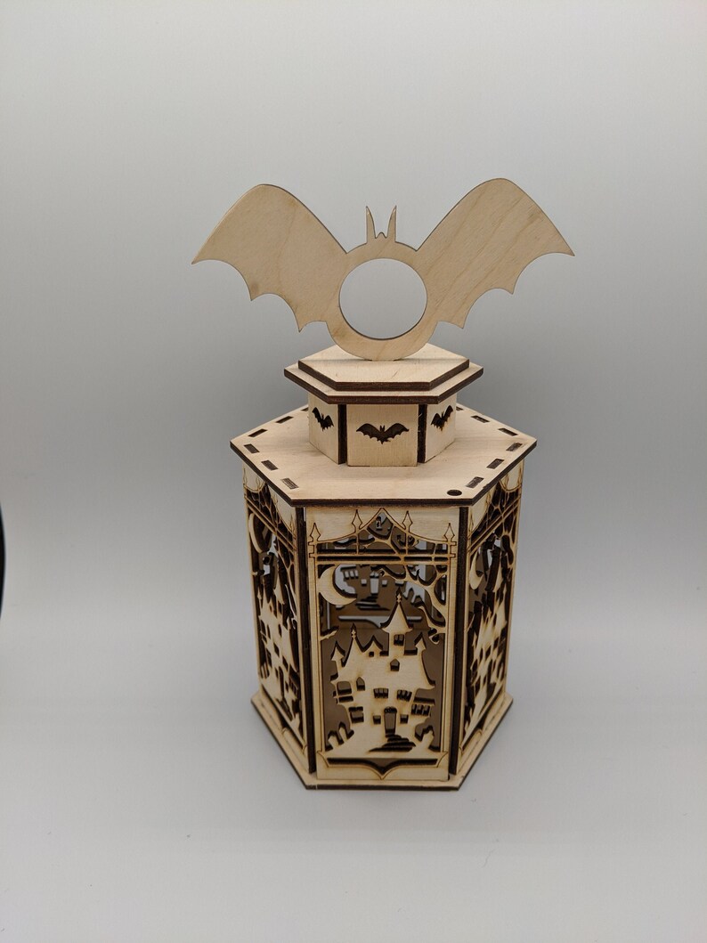 Peut inclure: Une lanterne d'Halloween en bois avec un motif de chauve-souris sur le dessus. La lanterne est en forme d'hexagone et pr&eacute;sente un motif d&eacute;coup&eacute; d'une maison hant&eacute;e avec une lune croissante.