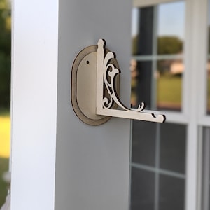 Puede incluir: Un soporte de estante de madera, montado en la pared, con una placa trasera ovalada y un estante en forma de L. El soporte presenta adornos decorativos y está hecho de madera de color claro. Está fijado a un pilar blanco.