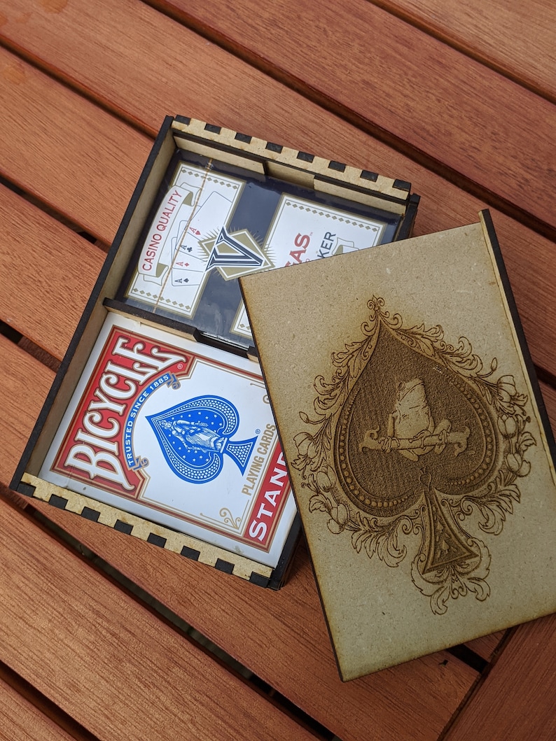 May include: A wooden box with a black and white chequered pattern on the inside. The box is open and contains a pack of Bicycle playing cards. The box is closed with a lid that has a brown background and a black and white design of a spade.