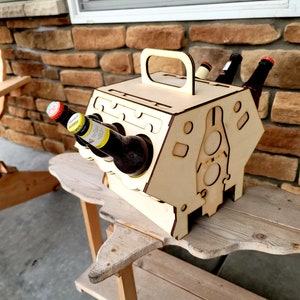Puede incluir: Portabotellas de madera con forma de bloque de motor. El portabotellas tiene un asa y tiene capacidad para seis botellas. El portabotellas está sobre una mesa de madera.