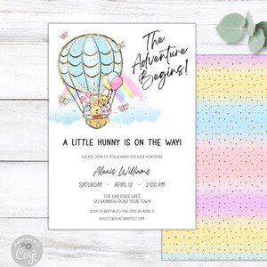 Könnte beinhalten: Eine Babyparty-Einladung mit einer Heißluftballon-Illustration. Der Ballon ist mit Winnie Puuh-Charakteren gefüllt und hat einen Regenbogen dahinter. Der Text lautet "The Adventure Begins!" und "A Little Hunny is on the Way!"