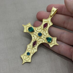 May include: A gold-coloured cross pendant with intricate filigree detailing. The cross features emerald green, ruby red, and sapphire blue gemstones. The pendant is held in a hand against a neutral background.