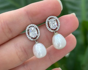 May include: A pair of silver drop earrings. Each earring has a clear oval-cut stone surrounded by smaller clear stones. A white, teardrop-shaped pearl hangs below each stone setting. The earrings are held by a hand.