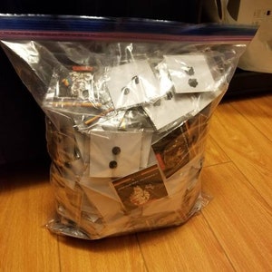 Könnte beinhalten: Ein klarer Plastikbeutel mit Reißverschluss, gefüllt mit verschiedenen kleinen Gegenständen, darunter Knöpfe und Karten. Der Beutel liegt auf einem Holzboden, und der Inhalt ist durch das transparente Material sichtbar.