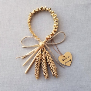 Peut inclure: Ornement artisanal en blé et paille. Il présente un anneau en paille tressée, des épis de blé et une petite étiquette en forme de cœur en bois avec du texte. Le design est noué avec de la ficelle, pour une esthétique naturelle.