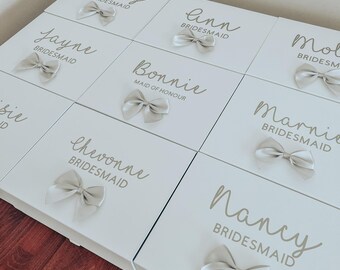 May include: White gift boxes with gold lettering and a satin bow on each box. The boxes are labeled with names and titles such as "Maid of Honor", "Bridesmaid", and "Becky".