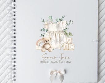 May include: A white spiral-bound baby memory book with a watercolor design. The cover features a baby onesie, teddy bear, rainbow, and ABC blocks. The name "Sarah Jane" and birth details are printed below the illustrations. A white bow is attached.