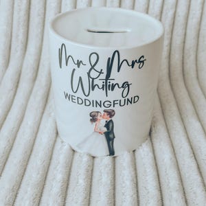 May include: A white ceramic wedding fund bank with a slot on top. The bank features the text "Mr & Mrs Whiting WEDDINGFUND" in black script. A graphic of a kissing bride and groom is below the text.