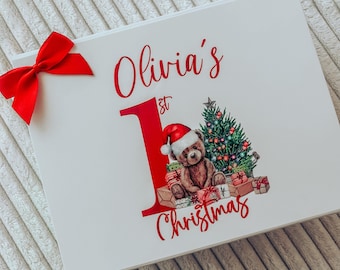 Könnte beinhalten: Eine weiße rechteckige Geschenkbox mit einer leuchtend roten Satinschleife auf der linken Seite. Der Text lautet Olivia's 1st Christmas in roter Schrift, ergänzt durch eine Grafik eines Teddybären mit Weihnachtsmütze neben einem Baum.