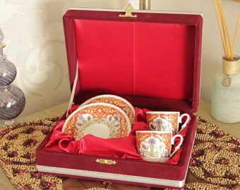 May include: A red velvet box with a gold trim and a gold clasp, containing two white porcelain teacups and saucers with floral designs. The box is sitting on a gold and red beaded table runner.