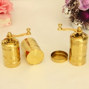 May include: Two gold metal pepper grinders with a crank handle. The grinders are on a white surface.