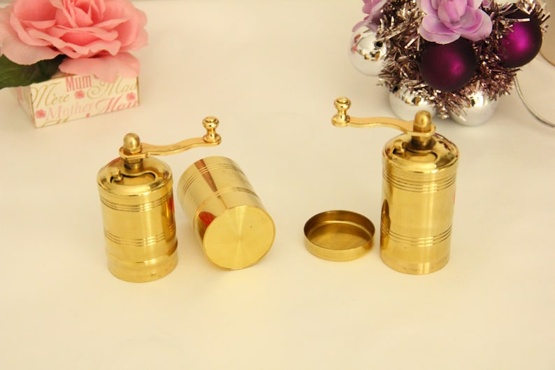 May include: Two gold metal pepper grinders with a crank handle. The grinders are on a white surface.