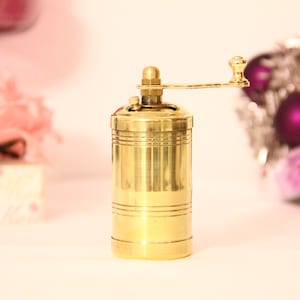 May include: A gold-colored brass manual pepper grinder with a crank handle. The grinder has a cylindrical body with subtle horizontal lines, and a small knob on top. It's a kitchen utensil used for grinding spices. The background is blurred, showing pink roses and a decorative item. Pepper grinder. Manual mill. Manual grinder. Manual pepper mill.