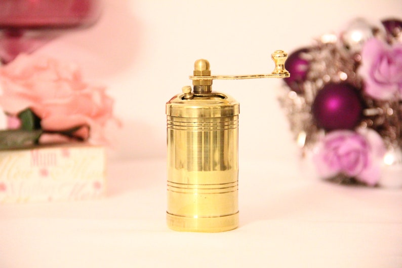 May include: A gold-colored brass manual pepper grinder with a crank handle. The grinder has a cylindrical body with subtle horizontal lines, and a small knob on top. It's a kitchen utensil used for grinding spices. The background is blurred, showing pink roses and a decorative item. Pepper grinder. Manual mill. Manual grinder. Manual pepper mill.